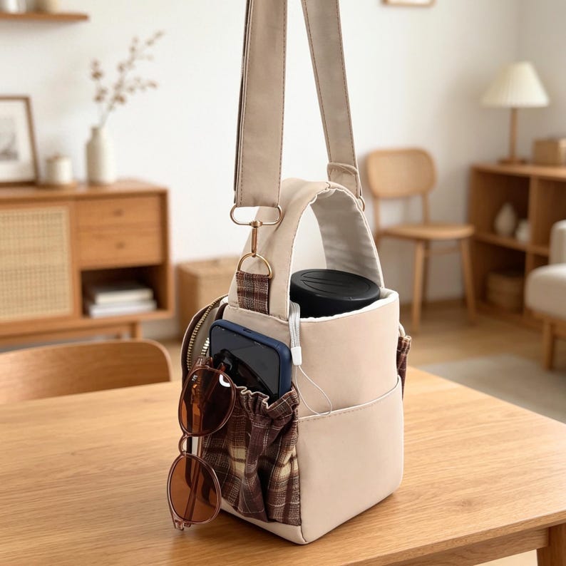 May include: A beige crossbody bag with a top handle and an adjustable strap. The bag features an exterior pocket with a brown and beige plaid pattern. A dark blue smartphone and brown-tinted sunglasses are tucked into the plaid pocket.