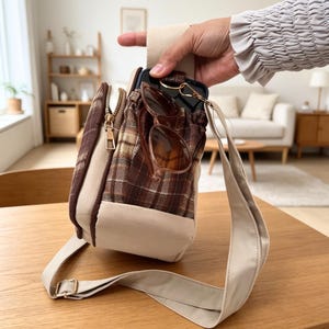 May include: A brown and beige plaid crossbody bag featuring a cream-colored base and strap, a gold-tone zipper, and a front pocket containing a black smartphone and brown-tinted sunglasses. The bag has a structured rectangular shape.
