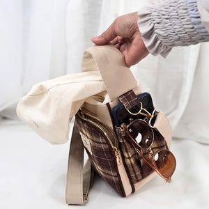 May include: A brown and beige plaid crossbody bag featuring a solid beige strap, a front zippered pocket, and an open side pocket containing a dark smartphone and brown-tinted sunglasses. The bag has a light beige fabric lining visible inside.