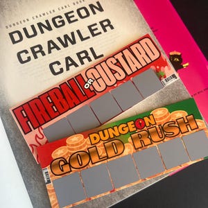 May include: Two rectangular scratch-off cards featuring the titles Fireball or Custard and Dungeon Gold Rush, each with five gray scratch-off panels, layered over a book cover titled Dungeon Crawler Carl Book One with a pink background.