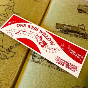May include: A rectangular white sticker with red graphics and text. It features the words ONE WISH WILLOW, AMAZE YOUR FRIENDS!, and You only get ONE WISH. The design includes illustrations of two faces, stars, hearts, and small icons.