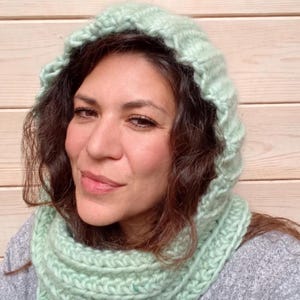 May include: A light mint green chunky knit hooded scarf featuring a thick ribbed texture. The accessory consists of a deep hood that transitions into a long, wrap-around scarf section, crafted from soft, thick yarn with a consistent stitch pattern.