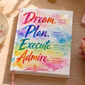 May include: A white journal with the words "Dream, Plan, Execute, Admire" in colorful lettering. The journal has a pink ribbon bookmark and is on a wooden surface. A cup of tea, flowers, and a notepad are also visible.