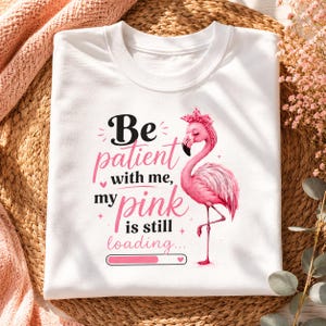 May include: White t-shirt featuring a pink flamingo illustration with a pink headband. The shirt has the text "Be patient with me, my pink is still loading..." in black and pink lettering. The design includes a loading bar.