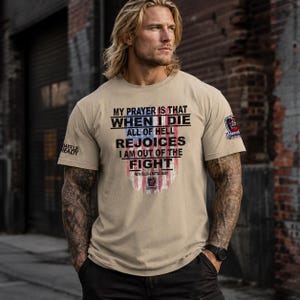 May include: Tan crew neck t-shirt with the text "MY PRAYER IS THAT WHEN I DIE ALL OF HELL REJOICES I AM OUT OF THE FIGHT". The shirt has a graphic of the American flag and the words "BATTLE READY" on the sleeve.