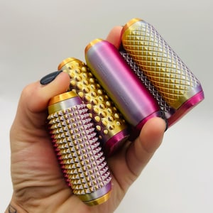 Könnte beinhalten: Eine Hand hält vier farbenfrohe, zylindrische Objekte. Die Objekte haben Gold-, Rosa- und Lilatöne. Zwei haben ein strukturiertes, genopptes Muster, und zwei haben ein Diamantmuster. Jedes hat goldene Akzente an den Enden.
