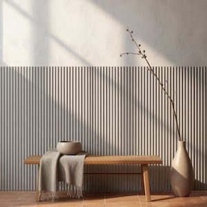 May include: A light wood bench with a beige throw blanket and a small bowl on top, positioned next to a tall beige vase containing a thin branch. The bench sits against a wall with vertical ribbed paneling and a smooth upper section.