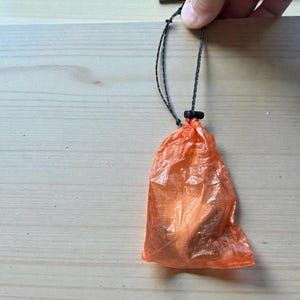 May include: An orange, translucent, drawstring bag with a black cord and toggle. The bag is held up by a black cord. The bag is made of a lightweight material.