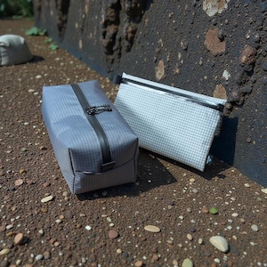 May include: Two travel pouches. One is a rectangular, gray pouch with a black strap and clasp. The other is a flat, white pouch with a grid pattern and a black zipper. Both are on a textured, brown surface.