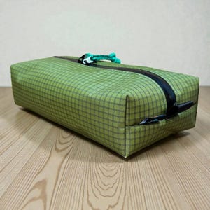 May include: A rectangular green travel pouch with a black zip and a green cord pull. The pouch has a grid pattern and is on a wooden surface.
