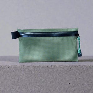 May include: A small, rectangular pouch with a green and white checked pattern. It has a black zip and a black fabric loop. The zip pull is a light green cord.