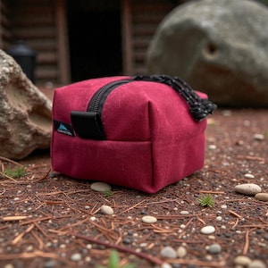 May include: A burgundy-colored, cube-shaped travel bag with a black zipper and a black handle. The bag is made of a textured fabric and has a small black tag with a logo on the side. A black and white rope is attached to the zipper.