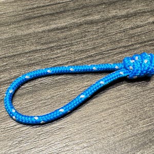 May include: A blue rope loop with white diamond-shaped accents and a knotted end. The rope is lying on a wooden surface. This item could be used as a keychain or a decorative element.