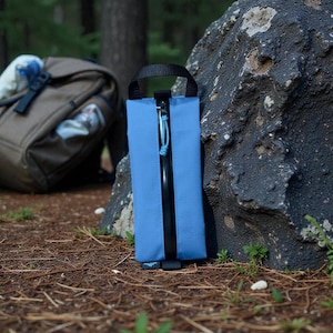 May include: A blue rectangular carrying case with a black zip and handle. The case is leaning against a large rock in a natural outdoor setting. A backpack is visible in the background.