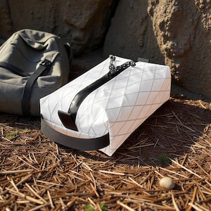May include: A white rectangular pouch featuring a grey diamond grid pattern, a black centre zip with a cord pull, and a black horizontal handle at the base. The pouch is constructed from a semi-translucent material with visible structural seams.