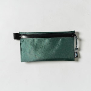 May include: A rectangular dark green zippered pouch featuring a diamond-grid pattern on the fabric. The pouch has a black zipper, a black pull tab, and a small black tag on the side with the white text WESTON and a mountain icon.