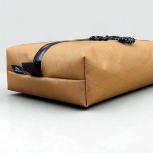 May include: A rectangular, tan-colored bag with a black zipper and a black clasp. The bag has a quilted pattern and a black accent on the side. The bag is sitting on a white surface.