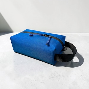 May include: A rectangular blue toiletry bag made of a durable woven fabric. The bag features a black zipper closure with two matching blue cord pulls and a sturdy black nylon webbing handle attached to the side for easy carrying.
