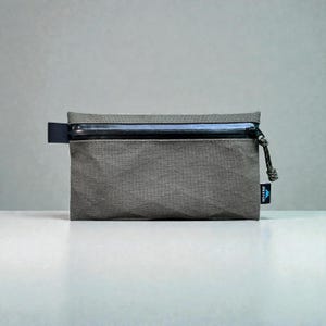 May include: A rectangular olive green zippered pouch with a textured, geometric surface pattern. It features a black horizontal zipper across the top, a black pull tab, and a small black fabric tag on the side with the white text WESTON.
