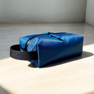 May include: A blue rectangular travel bag with a black zipper and a black handle. The bag is made of a textured material and has a blue zipper pull. The bag is sitting on a light-colored wooden surface.