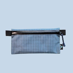 May include: A rectangular zipped pouch in a light blue grid pattern fabric. It features a black horizontal zip across the top, black side tabs, and a blue cord zip pull. A small black tag on the right side displays the text WESTON.
