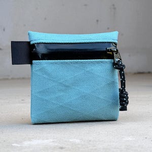 May include: A small, rectangular turquoise wallet with a black zipper and a black pull tab. The zipper pull is a black and white cord. The wallet is made of a textured fabric and sits on a concrete surface.