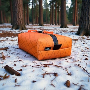 May include: An orange rectangular travel bag with a black strap and zipper closure, set against a snowy forest backdrop. The bag features a diamond pattern and the brand name "WESTON" on a small label.