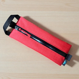 May include: A rectangular red pencil case featuring a subtle hexagonal pattern, a black water-resistant zipper with a red pull cord, and a black nylon webbing handle on one end. A small black fabric tag with white text is attached to the side.