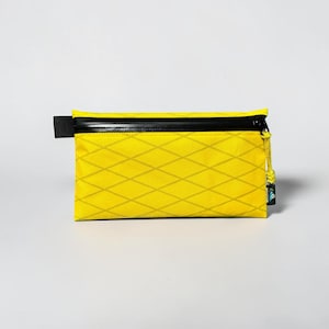 May include: A rectangular yellow zippered pouch featuring a diamond-shaped grid pattern across the surface. The pouch includes a black horizontal zipper closure along the top edge and a small yellow cord pull attached to the zipper slider.
