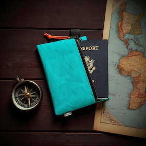May include: A teal passport holder with a diamond pattern, a black zip, and an orange pull cord. A compass and a vintage map are also visible on a dark wood surface. The word "SSPORT" is visible on the passport.