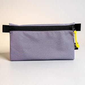 May include: A rectangular zippered pouch made of light purple fabric with a white grid pattern. It features a black horizontal zipper across the top, black side tabs, and a bright yellow cord pull attached to the zipper slider. A small black tag with white text is visible on the right side.