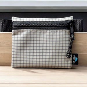 May include: A small, square pouch with a black zip and a black strap. The pouch is white with a black grid pattern. A black and white cord is attached to the zip. A small blue tag with white text reads "WESTON".