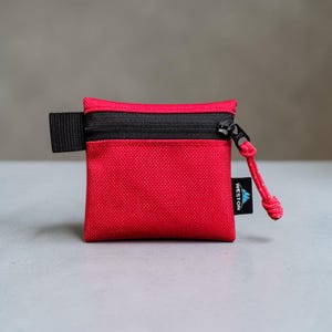 May include: A small, square, red zippered pouch made of a textured fabric. It features a black zipper across the top, a black side tab, and a black zipper pull with a red cord. A black tag on the side displays the white text WESTON with a mountain icon.