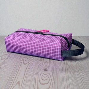 May include: A rectangular, purple toiletry bag with a white grid pattern. The bag has a black zipper, a pink zipper pull, and a black handle. The bag is sitting on a wooden surface.