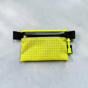 May include: A rectangular lime green pouch featuring a white grid pattern, a black horizontal zipper closure, and black side tabs. A bright neon yellow cord pull is attached to the zipper slider, and a small black fabric tag is on the right side.