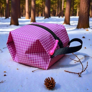 May include: A pink and white checkered travel bag with a black zipper and handle. The bag has a pink pull cord and is sitting on a snowy surface. A pine cone and twigs are also visible.