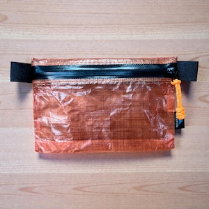 May include: An orange, rectangular pouch with a black zipper and strap. The pouch is made of a translucent material, with an orange pull cord and a black tag attached to the side.