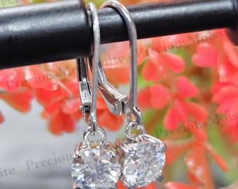 May include: A pair of leverback earrings with round, clear gemstones. The earrings are silver-toned and feature a leverback closure. The gemstones are set in a classic four-prong setting, and the earrings are displayed against a blurred background of red flowers.