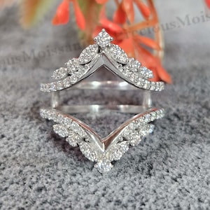 May include: A silver ring guard with a V-shaped design, adorned with numerous small, clear gemstones. The ring guard is designed to sit around a ring, enhancing its appearance. The gemstones sparkle against the silver setting.