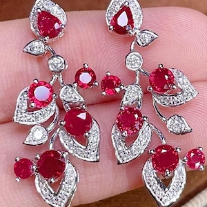 Peut inclure: Une paire de boucles d'oreilles pendantes au motif de branche en métal argenté. Chaque boucle est ornée de plusieurs rubis rouges ronds et en forme de poire, ainsi que de petites pierres claires pavées sur les éléments en forme de feuille.