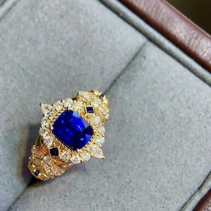 Peut inclure: Une bague en or ornée d'une grande pierre précieuse carrée bleu foncé au centre, entourée de petites pierres précieuses blanches et bleues. La bague présente un design orné et détaillé avec une bande texturée.