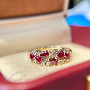 May include: A gold ring featuring a cluster of oval-cut, deep red gemstones, accented with smaller, clear gemstones. The ring is set in a gold band and is displayed in a red jewelry box.