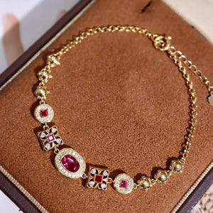 Peut inclure: Un bracelet en or orné de pierres précieuses rouges de formes variées, ovales, carrées et rondes, parsemées de petites pierres transparentes. Le bracelet est présenté sur un fond marron.