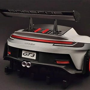 May include: A silver and black scale model of a Porsche GT3 RS sports car, viewed from the rear. The car features a large rear wing with the text PORSCHE, a red light bar, dual exhaust pipes, and a license plate clearly displaying the text GT3.