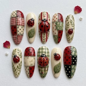 May include: A set of ten press-on nails featuring a variety of designs including red and white checkered patterns, polka dots, green leaf illustrations, and three-dimensional red ladybug embellishments on cream, red, and green backgrounds.