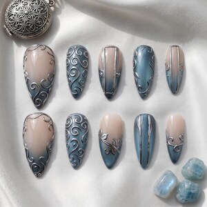 May include: A set of ten almond-shaped press-on nails featuring a gradient from nude to slate blue. The nails are decorated with raised silver filigree swirls, metallic vertical lines, and leaf motifs. A silver locket and three blue stones are included.