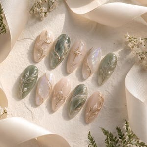 May include: A set of ten almond-shaped press-on nails featuring a palette of sage green, cream, and iridescent white. The designs include raised gold metallic leaf patterns, delicate pearl accents, and a central starburst motif on a cream base.