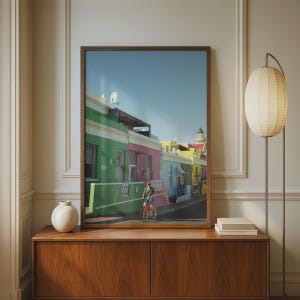 Può includere: Una stampa artistica da parete incorniciata che raffigura una scena di strada con edifici dai colori vivaci verde, rosa e giallo sotto un cielo azzurro. Una persona va in bicicletta lungo la strada asfaltata davanti alle case colorate con dettagli.