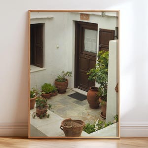 Peut inclure: Un miroir mural rectangulaire avec un cadre en bois clair reflétant une cour blanche. La scène montre une porte en bois brun foncé, un sol en pierre et plusieurs pots en terre cuite contenant des plantes vertes et de petits arbustes.