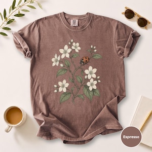 May include: Espresso-colored t-shirt featuring a floral and ladybug design. The graphic shows white flowers, green leaves, and a red and black ladybug. The shirt has rolled-up sleeves and a crew neck.